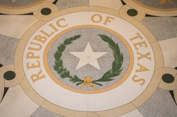 The Republic of Texas seal embedded in the floor of the Texas State Capitol building, featuring a lone star, olive and live oak branches, and gold lettering on terrazzo tile.